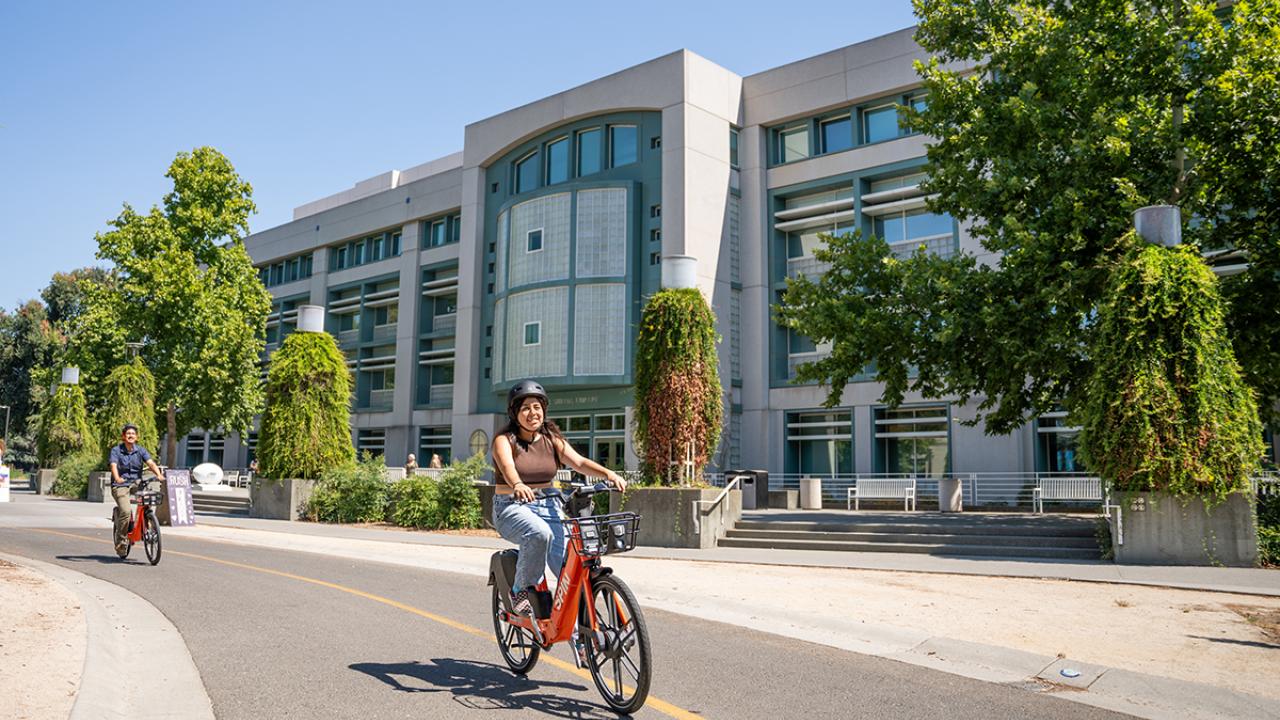 Image of student wearing a helmet, riding an electric bicycle in front of the UC Davis Library.