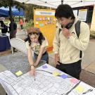 A young woman points to a place on a map while a young man looks on. They are at an outdoor tabling event about transporatation planning.
